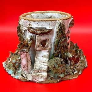Può includere: Un vaso di ceramica fatto a mano con un design unico e strutturato. Il vaso presenta una base bianca con accenti marroni e verdi, che ricorda un ceppo d'albero in miniatura con una piccola casa e dettagli di fogliame. L'interno è non smaltato.