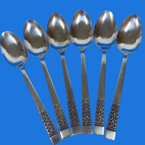 May include: A set of six silver-toned metal spoons arranged in a fan pattern. Each spoon features a smooth, polished oval bowl and a flat handle decorated with a repeating embossed scrollwork pattern near the base, ending in a straight edge.