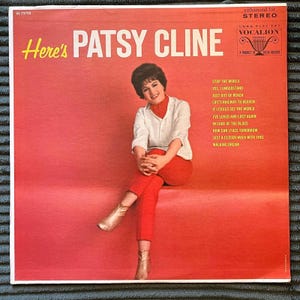 Peut inclure: Une pochette de disque vinyle vintage intitulée Here's Patsy Cline, montrant une photographie d'une personne aux cheveux bruns portant une chemise blanche et un pantalon rouge. Le fond est rouge uni, avec une liste de titres imprimée en jaune.
