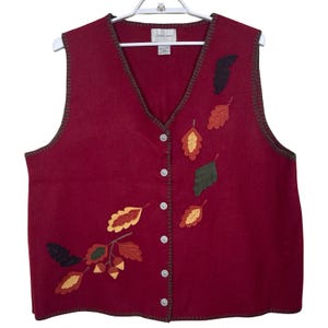 Puede incluir: Un chaleco burdeos con cuello en V y cierre de botones. El chaleco está decorado con hojas aplicadas en tonos de rojo, naranja, amarillo y verde. El chaleco está colgado de una percha blanca.