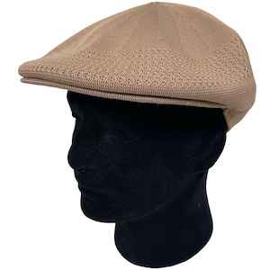 May include: A tan-colored newsboy cap with a rounded crown and a short, stiff brim. The cap is made of a knitted material, and it is displayed on a black mannequin head. The cap's texture is visible, and the color is consistent throughout.