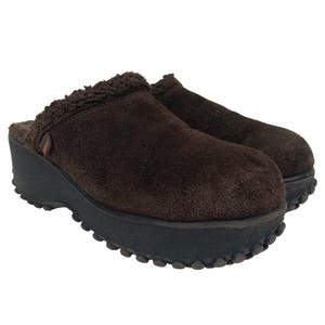 May include: A pair of brown suede clogs with a black platform sole. The clogs have a faux fur lining and a small logo detail on the side. The soles have a textured, rounded design.