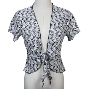 May include: A short-sleeved bolero cardigan made of white lace with a black leaf-like pattern. The garment features an open front design that ties at the waist with two fabric ends, creating a knot. The edges are finished with a thin black trim.