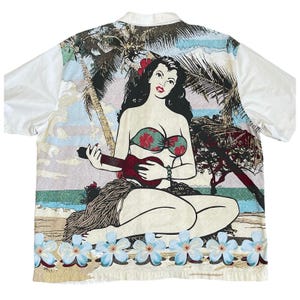 May include: White short-sleeved shirt with a colorful Hawaiian-themed graphic on the back. The graphic features a woman playing a ukulele on a beach, with palm trees and flowers. The shirt has a classic button-down design.