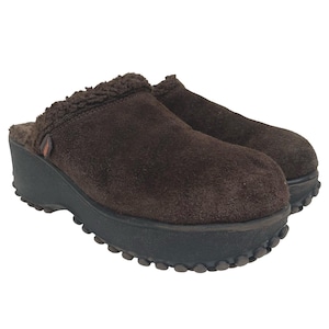 May include: A pair of dark brown suede clogs featuring a thick black platform sole with a textured, nubbed tread. The interior is lined with dark brown faux shearling, and a small red and white tag is visible on the side of the upper material.