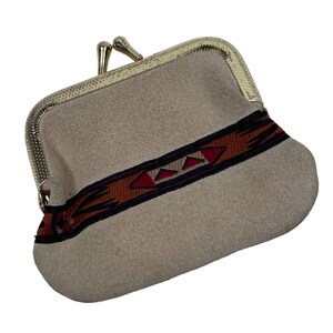 May include: A beige suede coin purse featuring a gold-toned metal kiss-lock frame. A horizontal band of woven fabric with a geometric pattern in shades of rust, black, and cream runs across the center of the purse, adding a decorative detail.