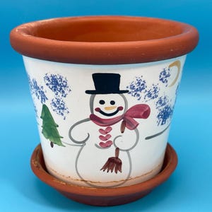 Könnte beinhalten: Weißer Keramik-Blumentopf mit Schneemann-Design. Der Schneemann hat einen schwarzen Zylinder, einen roten Schal und einen Besen. Der Topf hat einen Terrakotta-Rand und steht auf einer passenden Untertasse. Der Hintergrund ist blau.