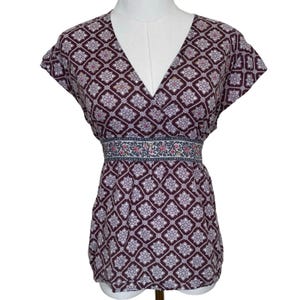 May include: A short-sleeved top with a V-neck and a geometric pattern in shades of burgundy, lavender, and white. The waistline features a decorative band with floral and leaf motifs. The top is displayed on a mannequin.