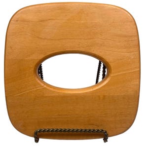 May include: A square light brown wooden tissue box cover with rounded corners and a smooth finish. The centre features a large, horizontal oval-shaped cutout for dispensing tissues. The item is supported by a dark, twisted metal wire stand.