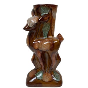 Könnte beinhalten: Eine Keramikvase in Form eines stehenden Hirsches mit glänzender brauner Glasur und strukturierten cremefarbenen sowie grünen Akzenten an Kopf, Rücken und Basis. Der Hirsch ist im Profil dargestellt, wobei der leicht gedrehte Kopf den Gefäßkörper bildet.