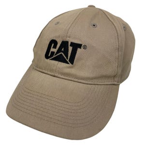 Peut inclure: Une casquette de baseball beige avec le texte noir brodé CAT accompagné d'un élément de logo triangulaire. La casquette présente une calotte structurée, une visière incurvée avec des coutures visibles et des œillets d'aération.