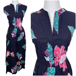 Könnte beinhalten: Ein ärmelloses, marineblaues Midikleid mit einem lebendigen tropischen Druck aus rosa Hibiskusblüten und grünen Palmblättern. Das Kleid hat einen geschlitzten V-Ausschnitt, ein eng anliegendes Oberteil und eine geraffte Taille mit einem Faltenrock.