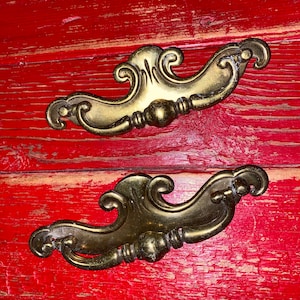 Può includere: Due tiretti per cassetti dorati decorati con un motivo a volute sono montati su una superficie in legno verniciata di rosso.