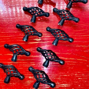 May include: Ten black metal cabinet knobs with a twisted, cage-like design. The knobs are arranged in a scattered pattern on a red wooden surface.