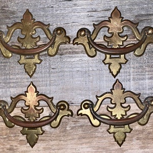 May include: Three ornate gold-toned drawer pulls with intricate designs. Each pull has a curved handle and a decorative, floral-inspired design.