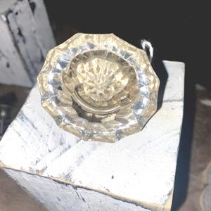 Pode incluir: Um puxador de porta de vidro transparente e facetado com um centro decorativo em forma de estrela. O puxador é octogonal e tem um pequeno fio branco preso. O puxador da porta está apoiado em um bloco de madeira pintado de cinza e branco desgastado.