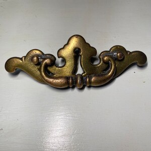 Op de afbeelding: Een gouden metalen kastgreep met een decoratief ontwerp en een sleutelgat in het midden.