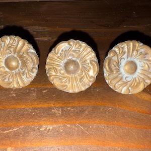 Puede incluir: Tres tiradores de armario dorados antiguos con un diseño floral. Cada tirador tiene un elemento circular central rodeado de patrones ornamentados y arremolinados. Los tiradores están colocados sobre una superficie de madera.