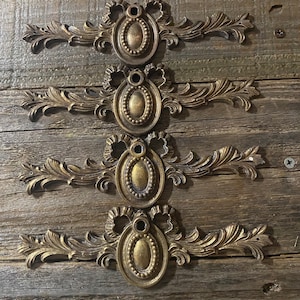 Puede incluir: Cuatro tiradores de cajón de metal dorado adornado con intrincados diseños florales y centros ovalados. Los tiradores están dispuestos verticalmente sobre un fondo de madera rústica.