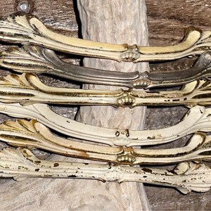 Pode incluir: Um conjunto de seis puxadores de gaveta antigos, ornamentados, brancos e dourados com um design floral decorativo.