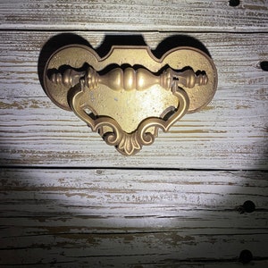 May include: A gold-toned metal drawer pull with a decorative, ornate design. The pull has a curved, heart-shaped base and a central handle with a rounded knob.