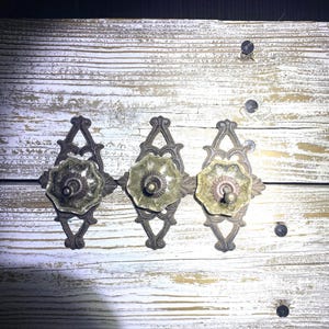 May include: Three antique-style hooks with ornate metal designs and clear glass knobs. The hooks are mounted on a whitewashed wooden surface.