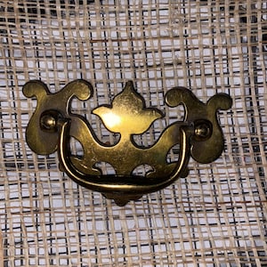 Könnte beinhalten: Ein goldfarbener Metall-Schubladenzug mit einem dekorativen, verzierten Design. Der Zug hat einen Schleifen-Griff und ist vor einem gewebten Hintergrund platziert.