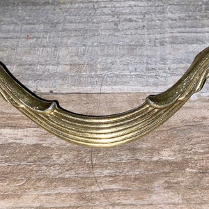 May include: A gold-toned metal drawer pull with a decorative, curved design. The pull has a fluted, ribbed texture and a scroll-like shape on each end.