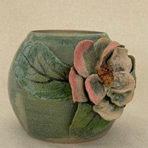 May include: A ceramic vase with a green glaze and a sculpted magnolia flower. The flower has pink and cream-coloured petals and a textured centre. The vase has a rounded shape and a painted leaf design.