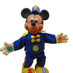 Könnte beinhalten: Ein Plüsch-Mickey-Mouse-Spielzeug, das eine blaue und gelbe Uniform mit einem roten, gelben, grünen und blauen Knopfdesign trägt. Das Spielzeug hat gelbe Schuhe mit roten, orangen, grünen und violetten Kreisen auf den Oberseiten.