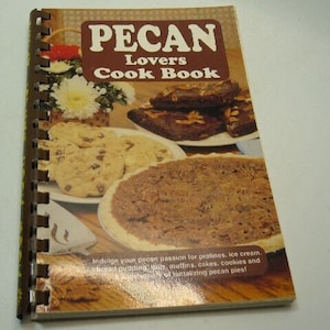 Op de afbeelding: Een bruin spiraalgebonden kookboek met de titel "PECAN Lovers Cook Book" in wit gedrukt. De omslag toont een foto van pecan-taart, pecan-koekjes en pecan-brownies.