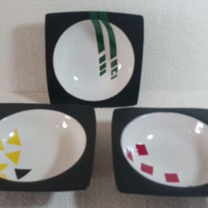 Peut inclure: Trois plats de service carrés avec un intérieur blanc et un extérieur noir. Chaque plat présente un design unique : des rayures vertes, des triangles jaunes et des carrés rouges. Ces plats décoratifs sont parfaits pour servir des collations ou des apéritifs.