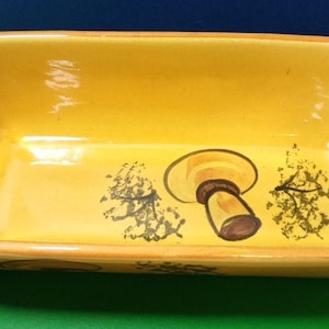 May include: A rectangular yellow ceramic baking dish with a mushroom design. The dish has rounded corners, handles, and a brown rim. The mushroom is brown and tan with a black outline.