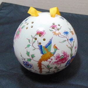 Puede incluir: Una esfera de cerámica blanca con un diseño floral y de pájaros. La esfera tiene una cinta amarilla atada en la parte superior y agujeros en toda la superficie.