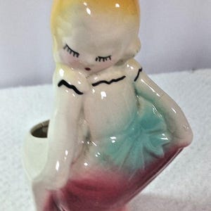 May include: A ceramic figurine planter shaped like a person with yellow hair, closed eyes, and a small red mouth. The figure wears a white top with black trim and a skirt featuring a teal bow and a deep pink base, with an open cavity at the back.