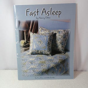 May include: A book cover titled "Fast Asleep" by Nancy Odom. The cover features a bed with blue floral pillows and a matching quilt. The book is about quilts, pillows, and totes for kids.