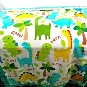 May include: A white fleece blanket featuring a colorful dinosaur pattern with green, blue, and yellow dinosaurs, palm trees, and the text "dinos" and "t-rex". The blanket has a teal satin border and is shown with a ruler measuring 36 inches.