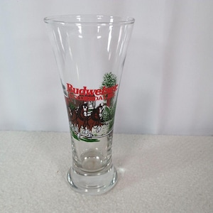 May include: Clear glass Budweiser beer glass with a flared top and a round base. The glass features the Budweiser logo in red and a graphic of horses pulling a carriage.