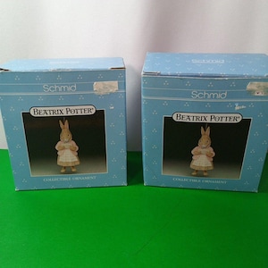 Puede incluir: Dos cajas azules con los nombres "Schmid" y "Beatrix Potter" impresos. Cada caja muestra un adorno coleccionable de un conejo con una chaqueta rosa y un vestido blanco. Las palabras "Collectible Ornament" están impresas debajo.