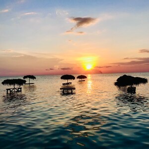 May include: Silhouettes of thatched umbrellas and tables in the ocean at sunset. The sun is setting over the horizon, casting a warm glow on the water.