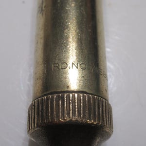 May include: Close-up of a brass-coloured metal object with the text "RD.NO.7156". The object has a textured, ridged base and a smooth, cylindrical body. The lighting highlights the metallic sheen.
