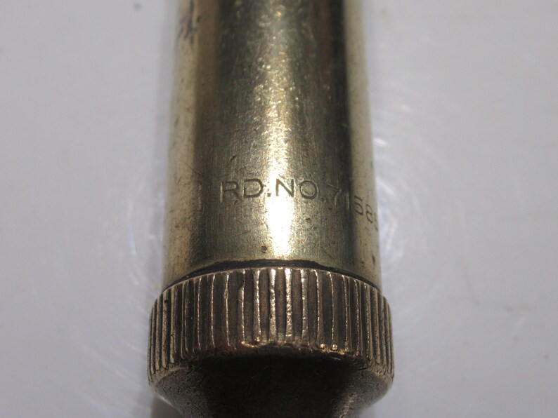 May include: Close-up of a brass-coloured metal object with the text "RD.NO.7156". The object has a textured, ridged base and a smooth, cylindrical body. The lighting highlights the metallic sheen.
