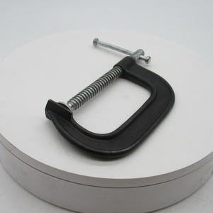 May include: A black C-clamp with a silver screw and threaded rod. The clamp is made of metal and is designed to hold objects together. The C-clamp is on a white surface.