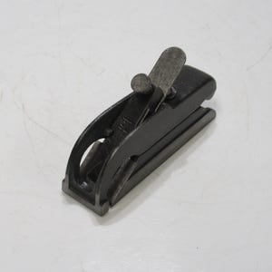 Puede incluir: Un pequeño cepillo de mano negro con una cuchilla y una tapa de palanca de metal. El cepillo tiene un cuerpo curvo y una suela plana. La tapa de la palanca está colocada en la parte superior y la cuchilla es visible en el centro. Herramienta de carpintería.
