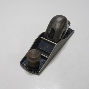 Puede incluir: Un cepillo de mano vintage, una herramienta para trabajar la madera, con un cuerpo de metal oscuro y un pomo y mango de madera. El cepillo tiene una base rectangular y un mango curvo. La herramienta está diseñada para alisar y dar forma a superficies de madera.