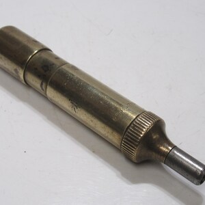 May include: A vintage brass tool with a cylindrical body and a tapered end. The tool has a tarnished appearance, suggesting age and use. The end is a darker metal. The tool is lying on a white surface.