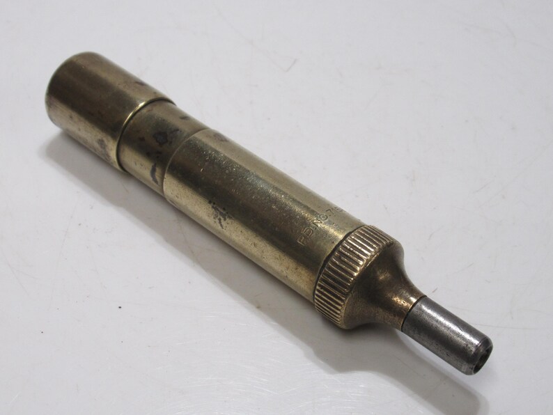 May include: A vintage brass tool with a cylindrical body and a tapered end. The tool has a tarnished appearance, suggesting age and use. The end is a darker metal. The tool is lying on a white surface.