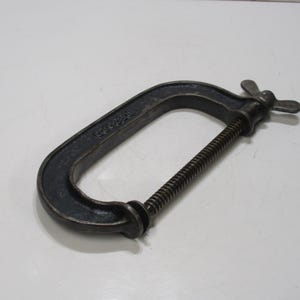 May include: A dark gray C-clamp, a tool used for holding workpieces together. The clamp has a C-shaped frame, a threaded spindle, and a butterfly handle for tightening. The tool is made of metal and sits on a white surface.