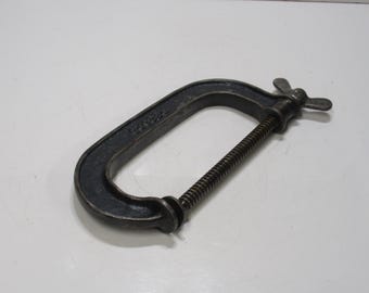 May include: A dark gray C-clamp, a tool used for holding workpieces together. The clamp has a C-shaped frame, a threaded spindle, and a butterfly handle for tightening. The tool is made of metal and sits on a white surface.