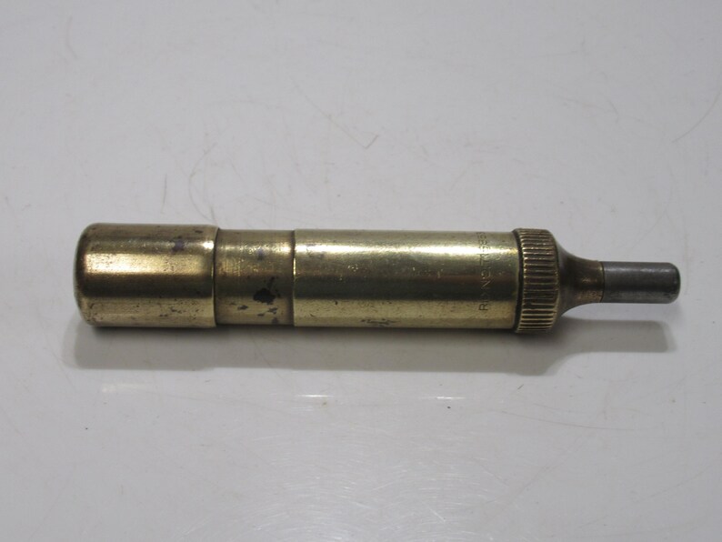 May include: A vintage brass object with a cylindrical body and a small, ridged end. The object has a tarnished, aged appearance, suggesting it is an antique. The end has a small, dark grey, cylindrical tip. The word "Rancaster" is printed on the side.